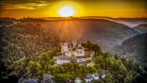 In einer hügeligen Mittelgebirgslandschaft steht im Fokus des Bildes eine große Burganlage auf einem Berg. Der Himmel ist orange und die Sonne ist nah über dem Horizont