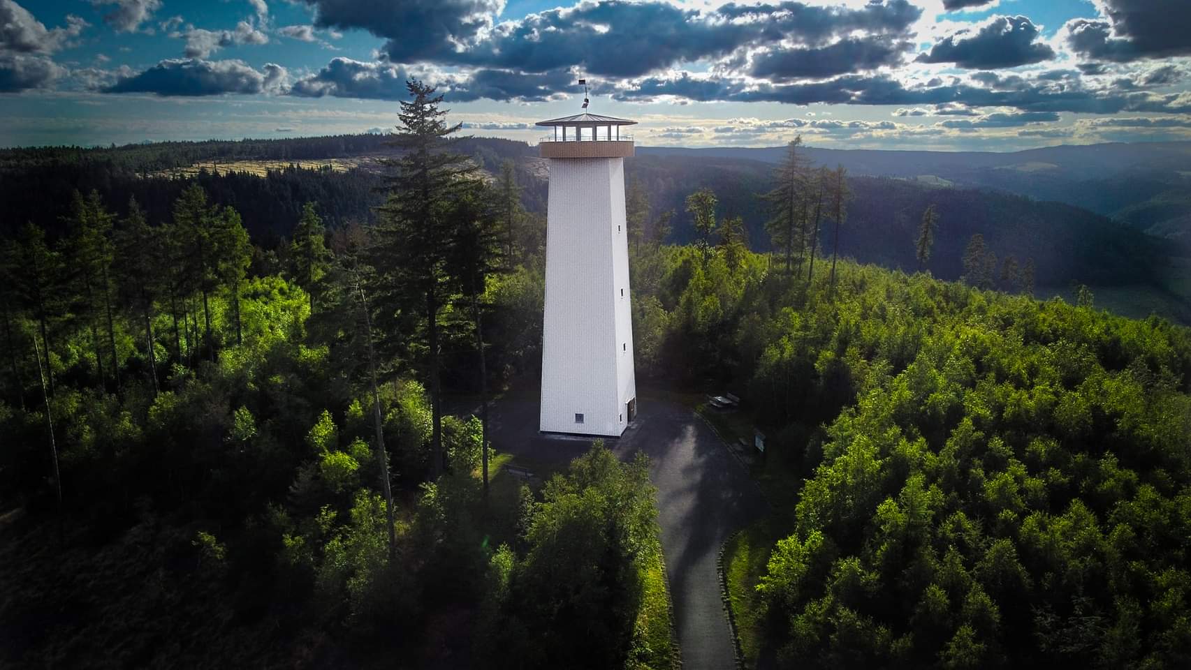 Fokus des Bildes ist ein weißer hocher Turm mit einer Aussichtsplattform. Er steht auf einem Hügel mit teilweise bewaldeten Flächen rundherum. Der Himmel ist mit dunklen Gewitterwolken verhangen.