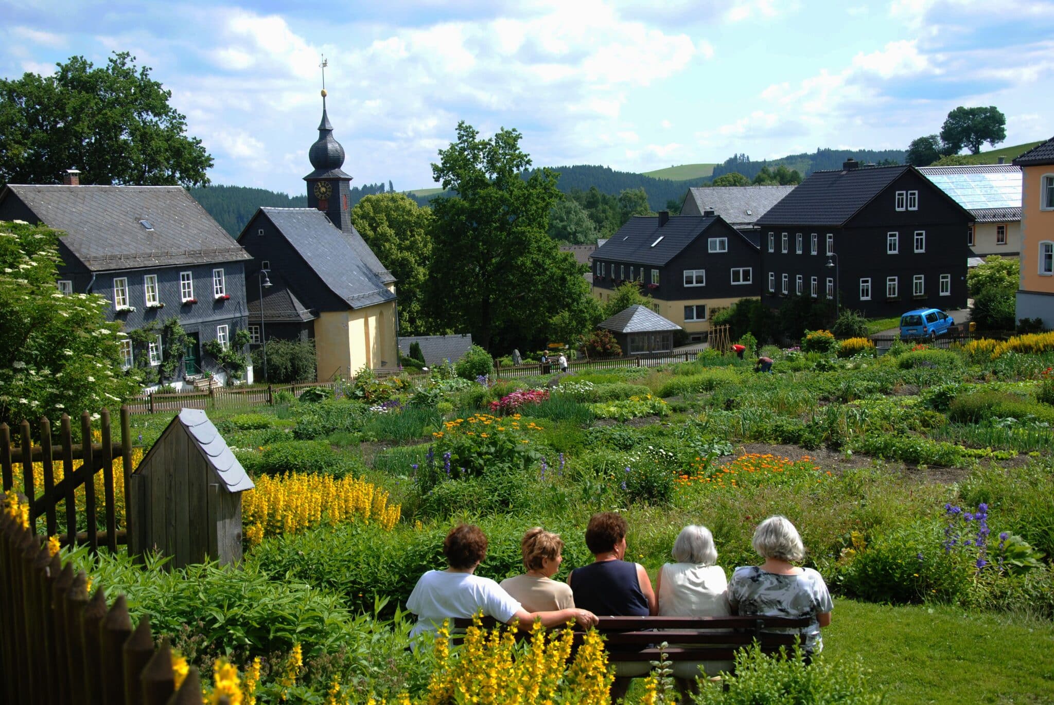 Eine Landschaftsaufnahme von einem großen Pflanzgarten mit Wohnhäusern und einer schiefergedeckten Kirche im Umfeld. Im Pflanzgarten sitzen Personen auf einer Bank.
