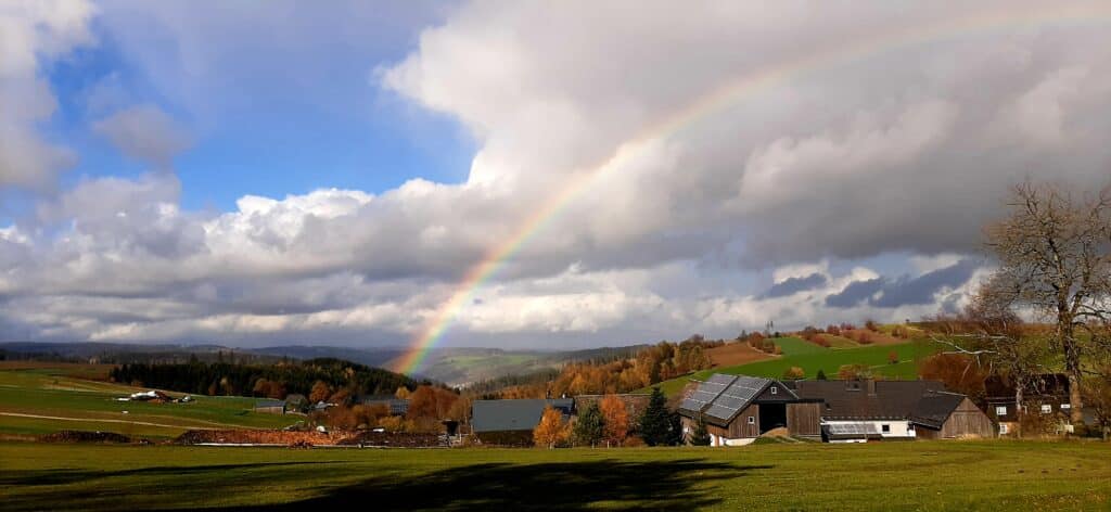 Eine Landschaft mit Wolken und teilweise blauem Himmel über einem Dorf. Ein großer Regenbogen ist über dem Dorf zu sehen.
