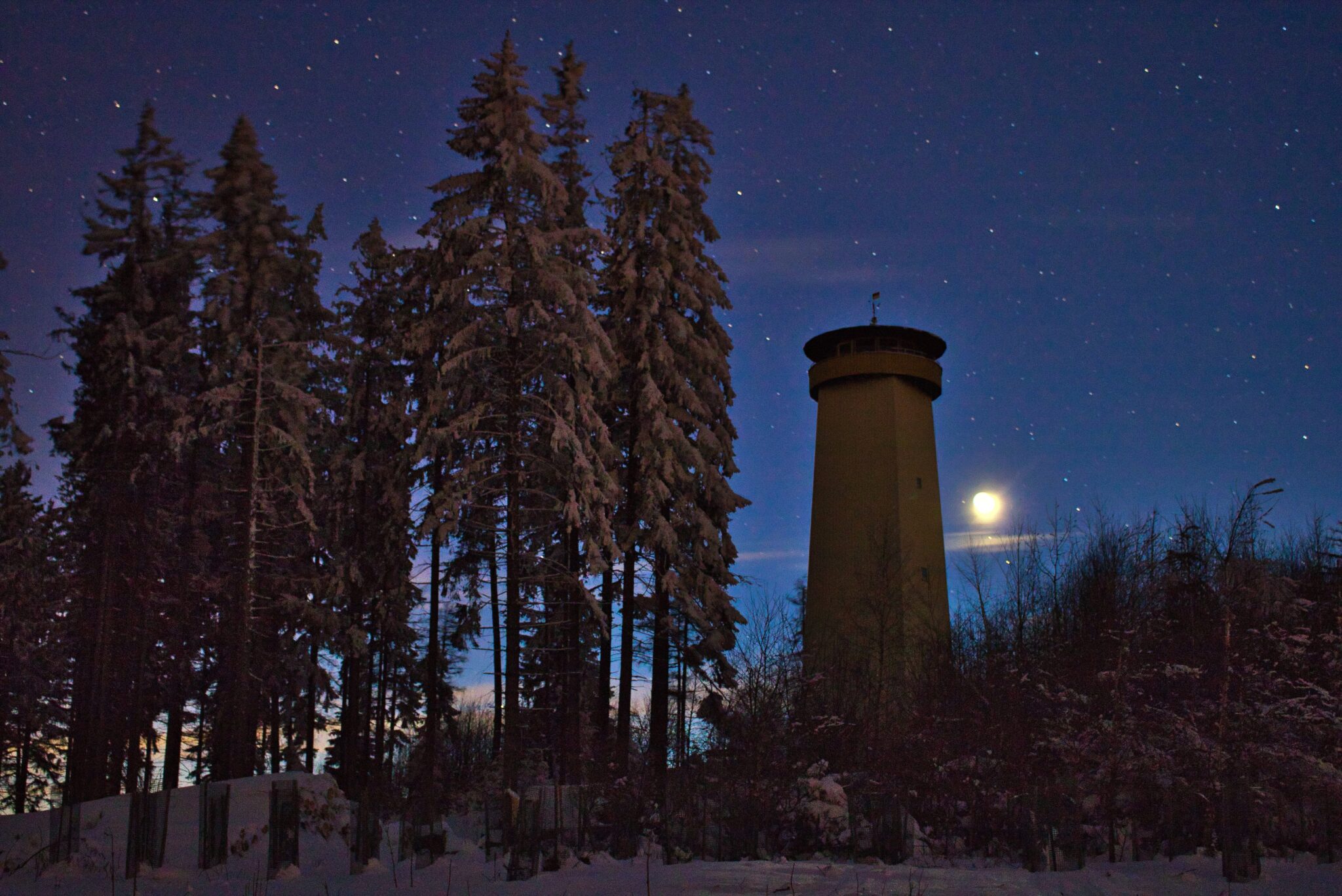 Nächtlicher Blick auf einen Aussichtsturm. Rundherum sind schneebedeckte Nadelbäume zu erkennen und es ist ein Vollmond neben dem Turm zu sehen.