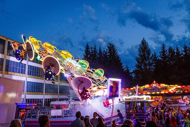 Zu sehen ist eine Volksfestszene mit einem Karussel in Bewegung und einem Festplatz mit abendlicher Beleuchtung